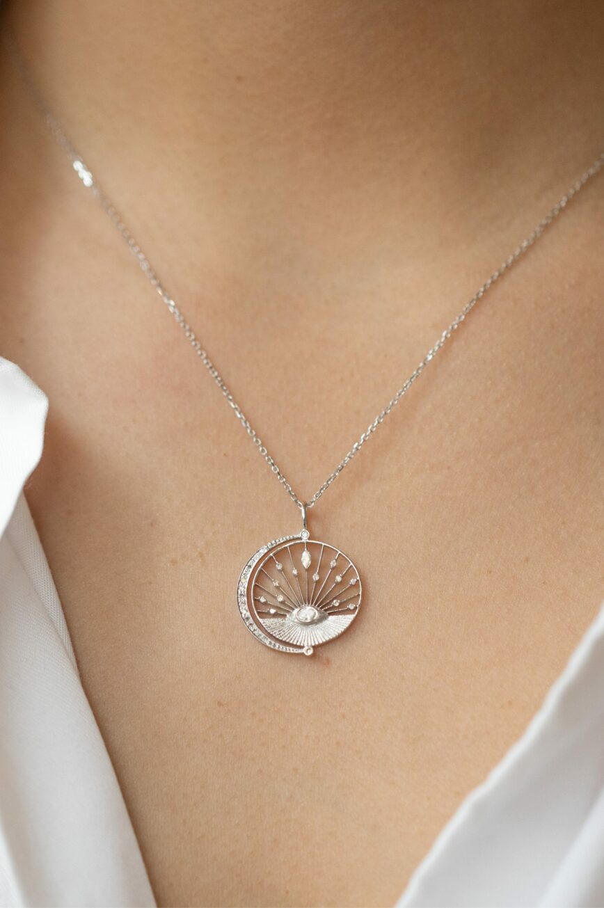 white gold circular medal necklace featuring an engraved sun eye motif at center with a crescent moon and pavé diamond accents, evoking a dreamlike celestial scene
