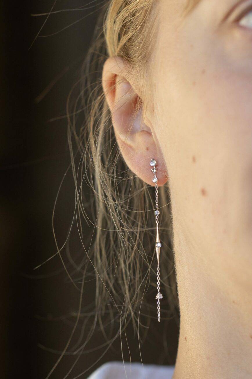 white gold long chain earrings each featuring a moonstone and diamond chain drop with dangling geometric details, worn as a delicate ear crawler