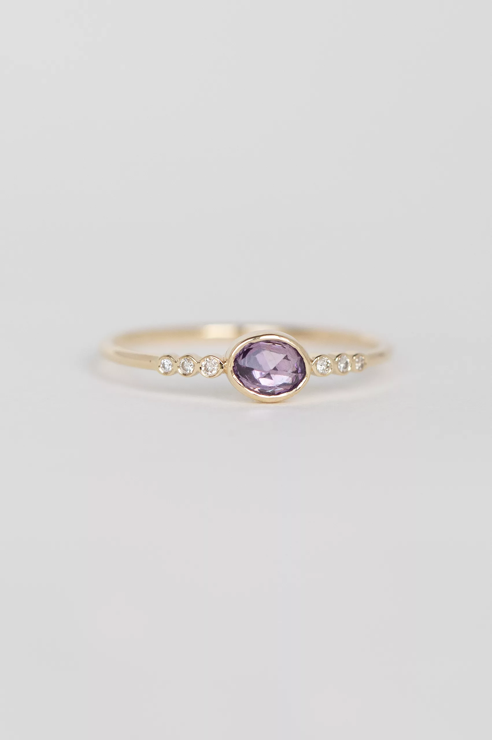 yellow gold ring with a bezel-set oval pink sapphire at center, flanked by a row of six round brilliant diamonds on a delicate band