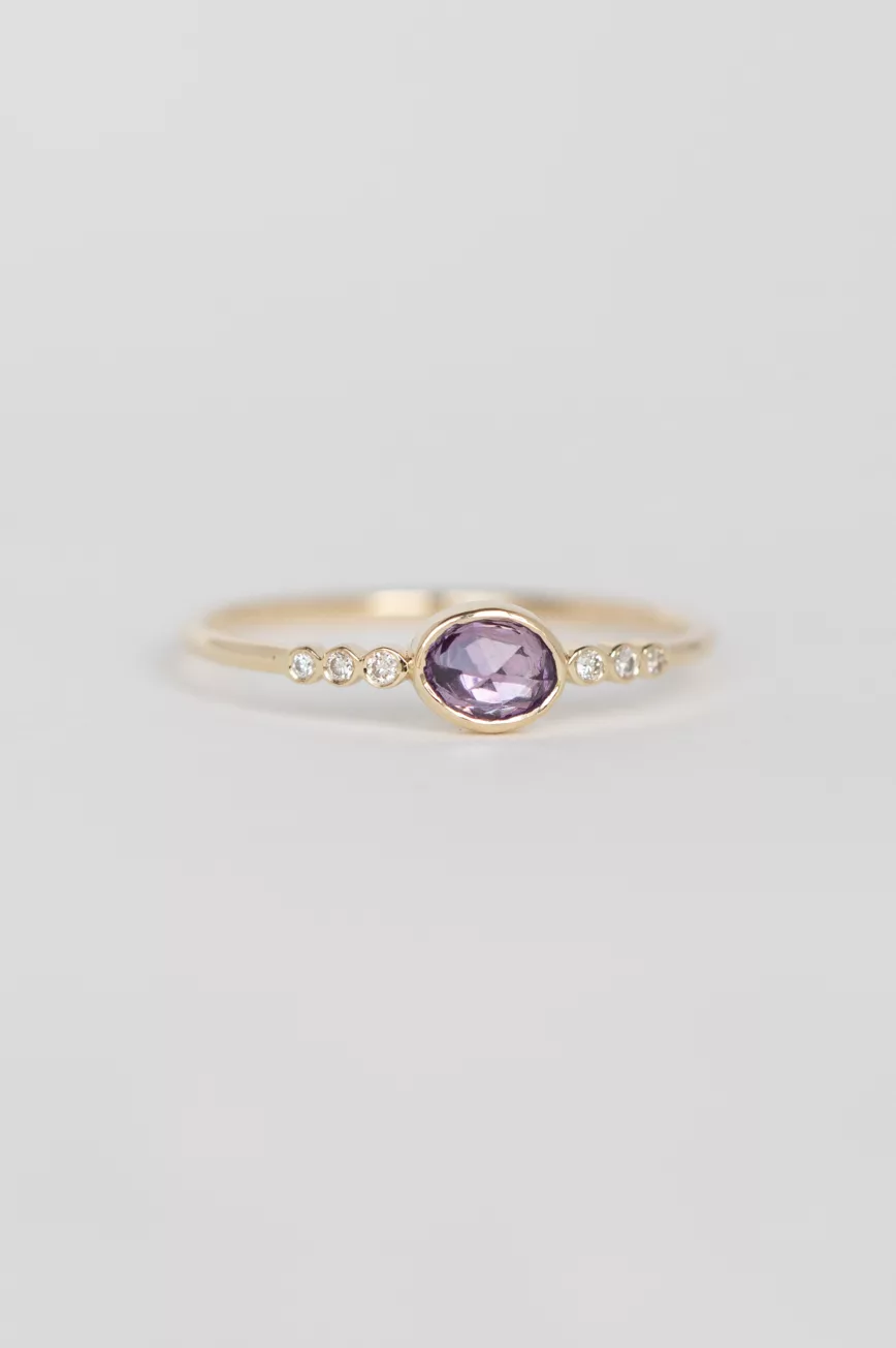 yellow gold ring with a bezel-set oval pink sapphire at center, flanked by a row of six round brilliant diamonds on a delicate band